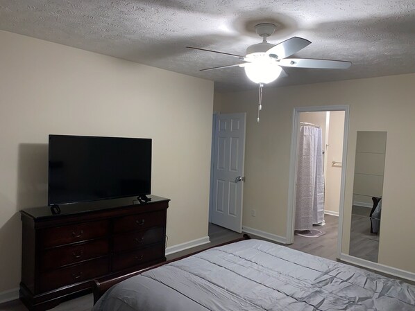 Un dormitorio con cama, cómoda, televisor, ventilador de techo y puerta que da a otra habitación.