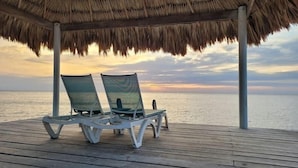Une terrasse en bois avec deux chaises longues, située sous un toit de chaume et offrant une vue sur la mer au coucher du soleil.
