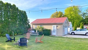 Una piccola casa con tetto rosso, un portico e un prato con due poltrone Adirondack e una panchina in legno.