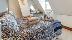 A bedroom with a sloped ceiling, a bed with floral bedding, a bedside table with a lamp, a mirror, and a window with a view. - Nans Cottage - UK43350 (Whitby)