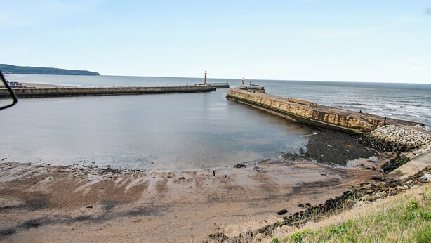 A coastal scene with a stone pier extending into calm waters, a lighthouse in the distance, and a sandy beach area. - Nans Cottage - UK43350 (Whitby)