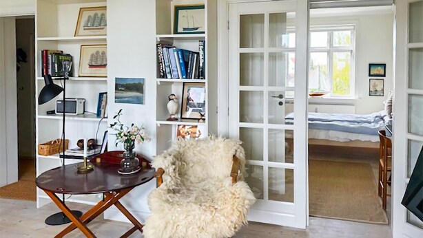 A cozy room with a wooden chair, a desk with a lamp, a bookshelf, and a bed with a window view.