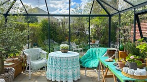 A conservatory with a tiled floor, a round table with a patterned tablecloth, a green sofa, and various potted plants.
