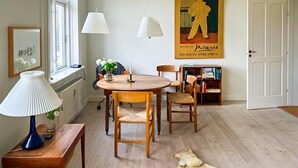 A dining area with a round wooden table, two chairs, a blue lamp, and a white lamp on a wooden side table. There is a poster on the wall and a bookshelf with books.