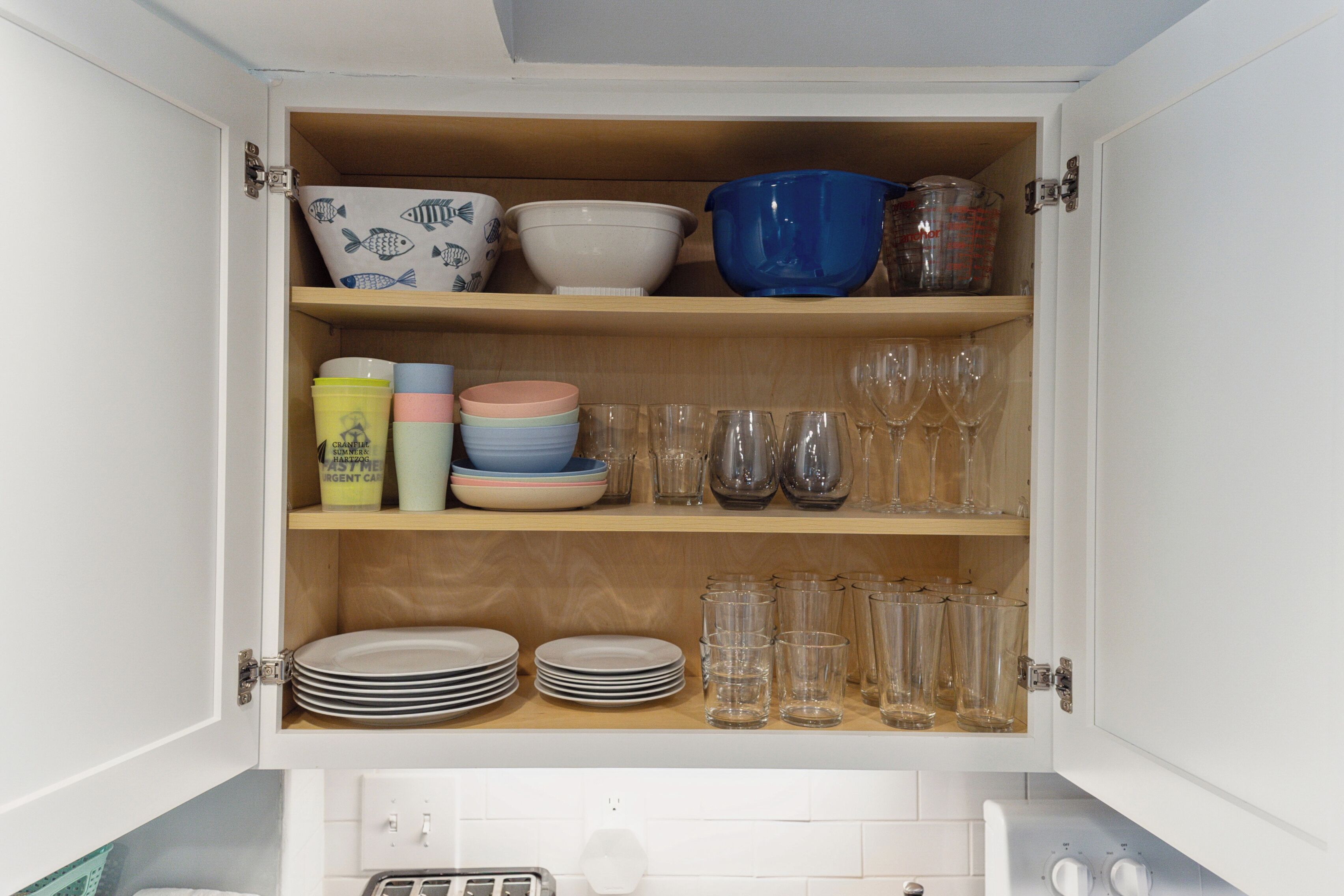 A kitchen cabinet with shelves containing dishes, bowls, glasses, and a coffee maker.