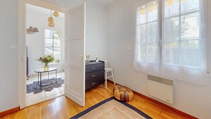 Una habitación con piso de madera, un aparador negro, una silla blanca, un otoman redondo y un ventanal con cortinas translúcidas.