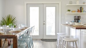 Een lichte keuken met een houten eettafel voor vier personen, witte barstoelen en een wit aanrechtblad met een koffieapparaat.