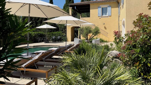 Area piscina con lettini e ombrelloni, immersa nel verde e affiancata da un edificio con tetto in tegole.