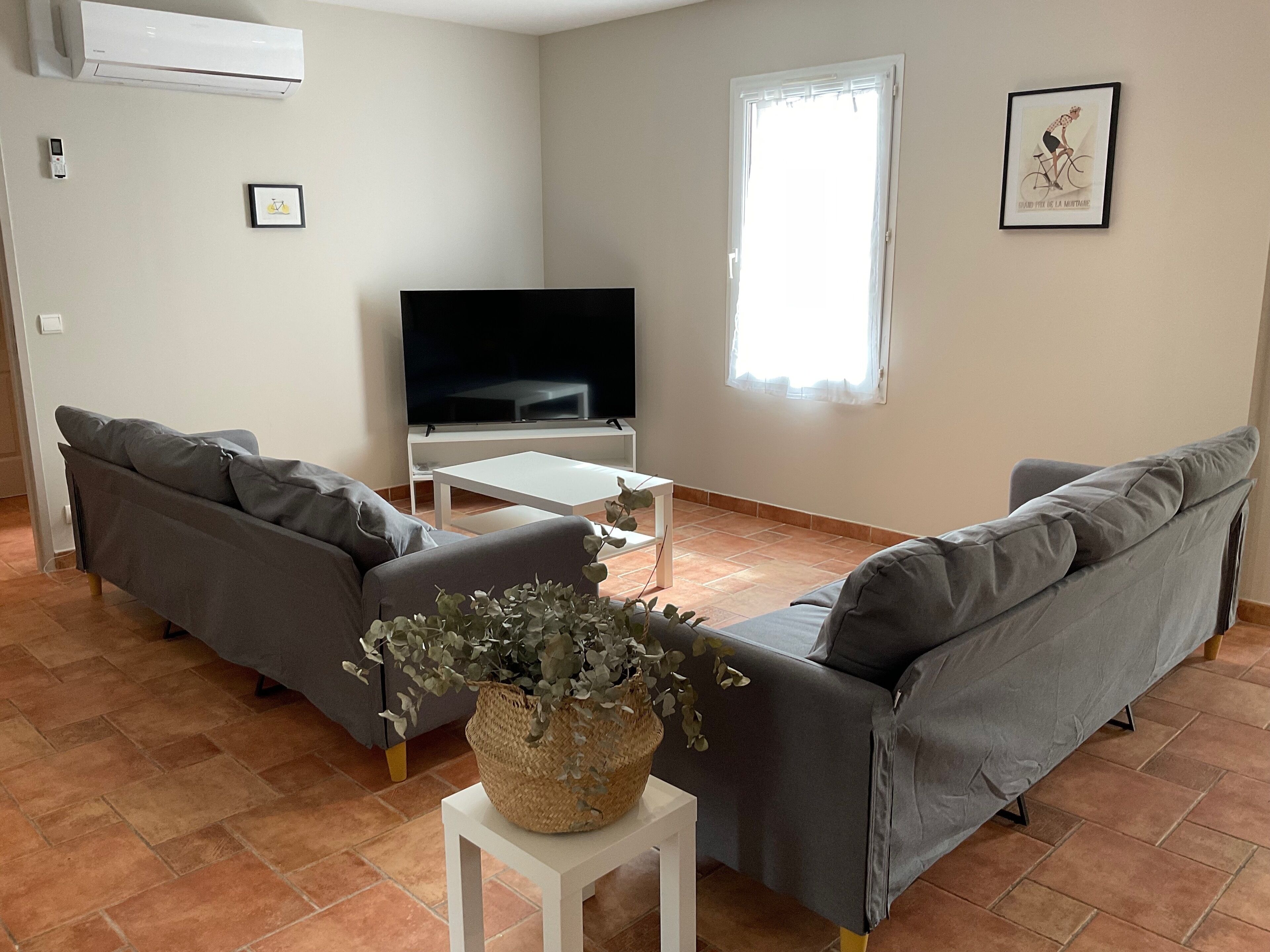 A living room with two grey sofas, a white coffee table, a flat-screen TV, a potted plant, and a window with curtains.