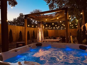 Un bain à remous agrémenté d’une pergola en bois et de guirlandes lumineuses, entouré d’une clôture en bois et d’arbres.