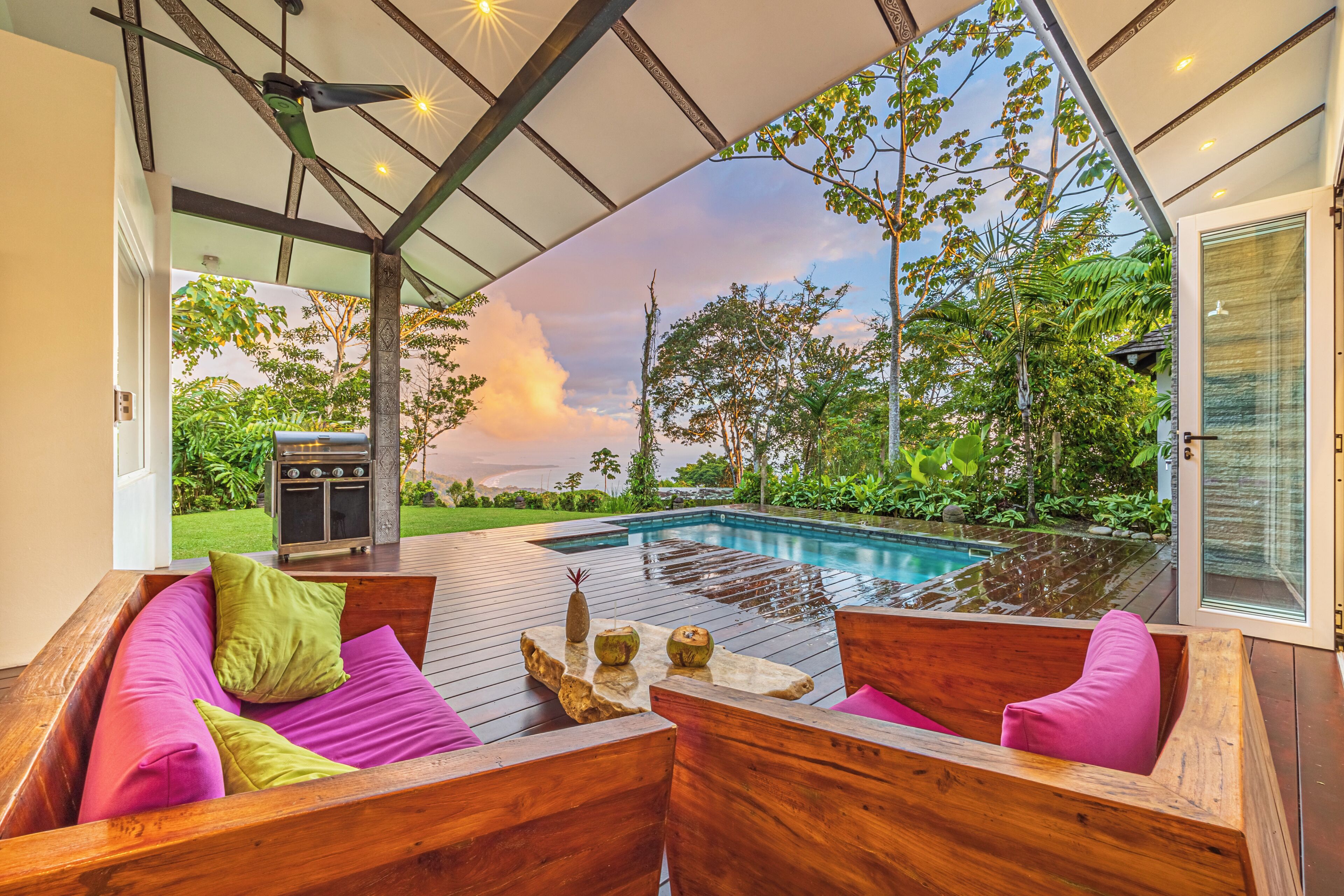 Una terraza de madera con piscina, mobiliario exterior y vistas al entorno verde.