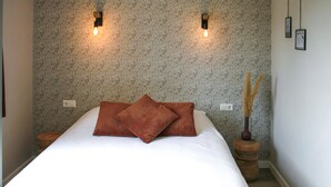 A neatly made bed with two pillows, a textured wallpaper, two wall-mounted lights, and a small wooden side table with a vase of dried plants.