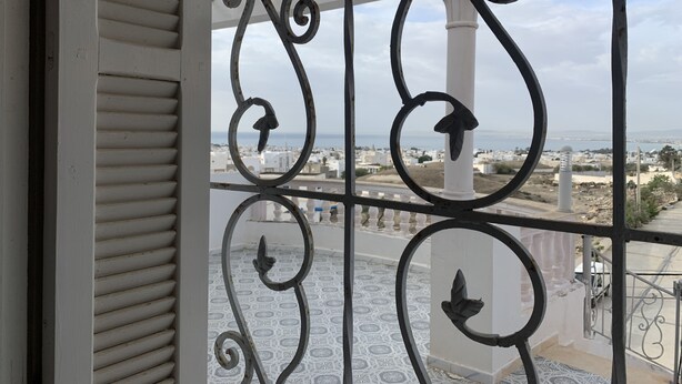 Un balcón con barandillas metálicas ornamentales y suelo estampado, con vistas a un pueblo costero de edificios blancos y el mar.