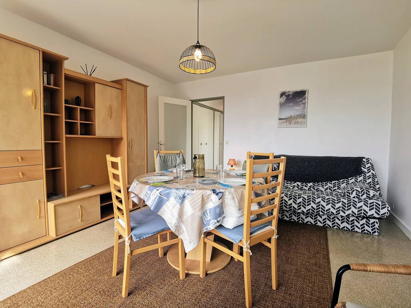 Un coin repas avec une table dressée pour deux personnes, des chaises en bois, un meuble avec des étagères et un luminaire suspendu.