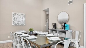Un comedor con una mesa rectangular dispuesta para seis personas, rodeada de sillas blancas. Una cafetera y un espejo están en la pared a la derecha.
