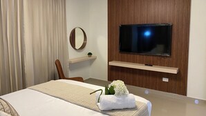 Une chambre d’hôtel avec un lit, un téléviseur fixé au mur, qui est recouvert d’un panneau en bois, une petite étagère avec une plante et un miroir.