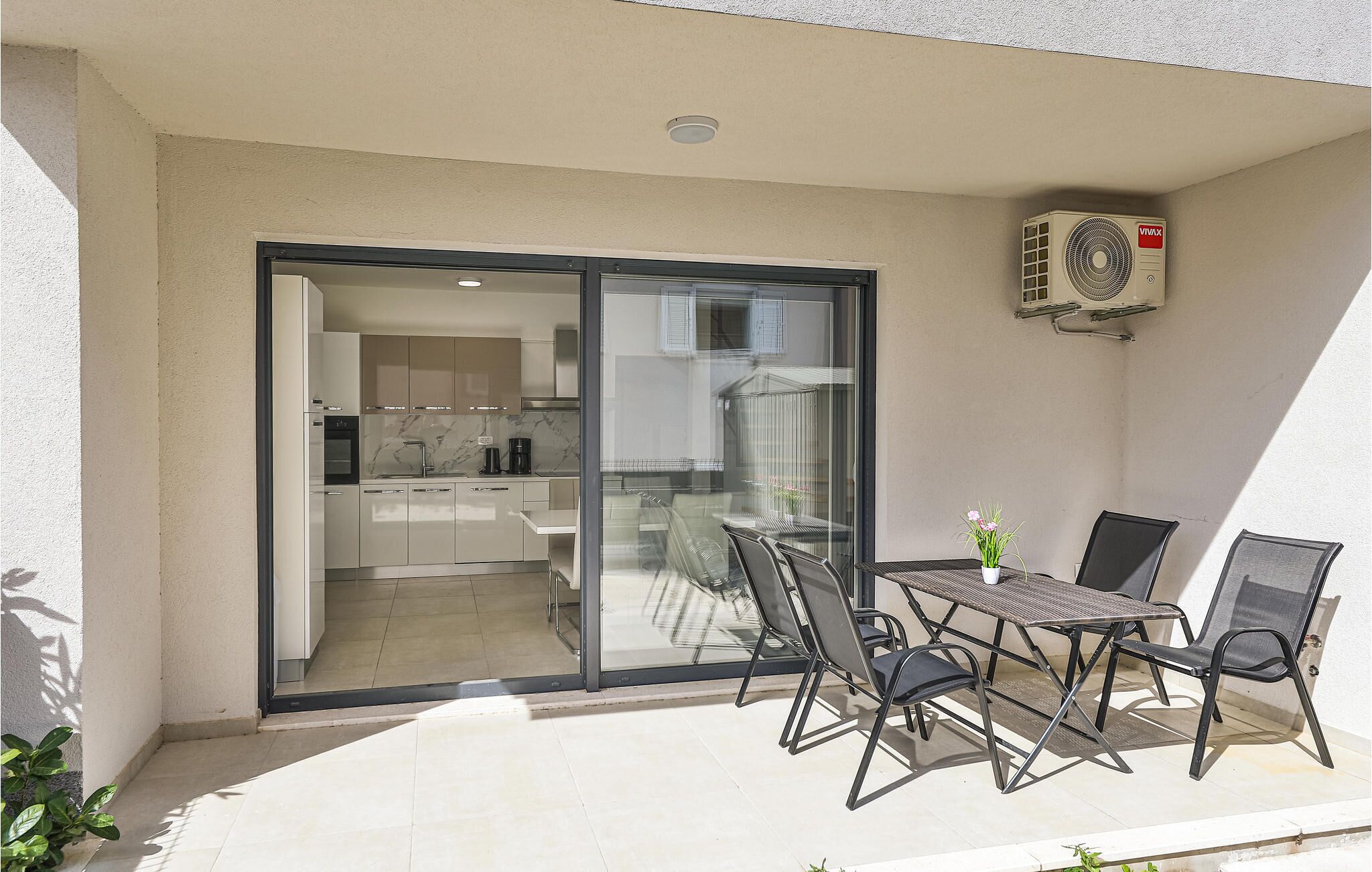 A balcony with a table and chairs, a sliding glass door leading to a kitchen, and an air conditioning unit on the wall.