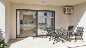 Uma varanda com mesa e cadeiras, uma porta de vidro deslizante que dá para uma cozinha, e um aparelho de ar condicionado na parede.