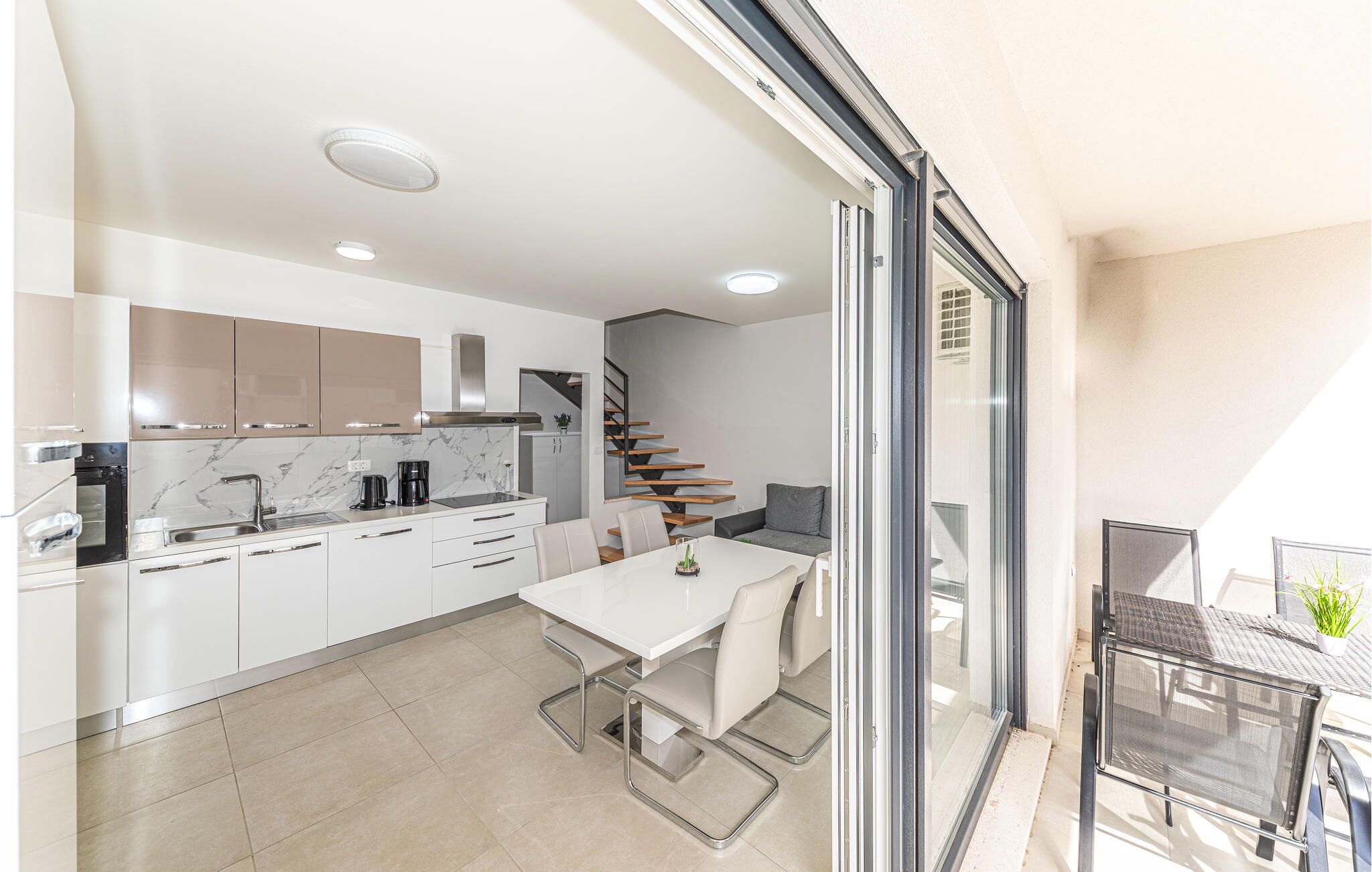 A modern kitchen with white cabinets, a marble backsplash, and a built-in oven. There is a dining area with a white table and chairs, and a staircase leading to an upper level.