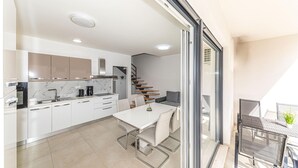 Cozinha moderna com armários brancos, painel de backsplash de mármore e forno embutido. Há uma área de jantar com mesa e cadeiras brancas, e uma escada que leva a um andar superior.