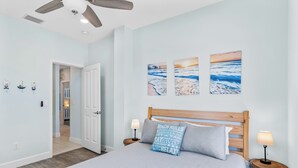 Een slaapkamer met een bed, twee nachtkastjes met lampen, een plafondventilator en wanddecoratie met strandtaferelen.