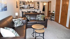 A living room with a brown leather sofa, two small round tables, and a dining area with a chandelier.