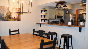 A dining area with a wooden table and chairs, a chandelier, and a kitchen area with shelves and appliances.