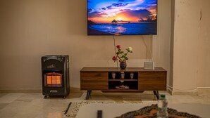 A living room with a flat-screen TV mounted on the wall displaying a sunset over water. A heater is on the floor to the left, and a wooden cabinet with a vase of flowers is in the center. - Modern & Large Studio in Casablanca - Bd Anfa (Casablanca)