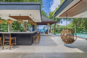 A modern outdoor bar area with seating and a hanging chair, overlooking a pool and lush greenery. - Casa Selva Ocean View Luxury Home- 4 Bedroom (Puntarenas Province)
