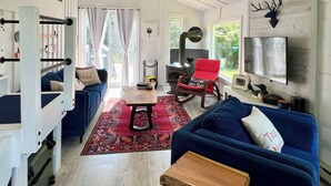 Een ruime woonkamer met een blauwe bank, een rode schommelstoel en een houten salontafel. Er is een open haard, een aan de muur bevestigde televisie en een groot raam met gordijnen.