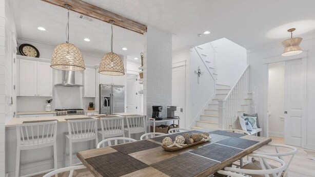 A modern kitchen with white cabinetry, a stainless steel refrigerator, and a dining area with a long wooden table and white chairs. - Longleaf Pines by AvantStay | Community Pool (Santa Rosa Beach)