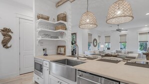 A modern kitchen with white cabinetry, a stainless steel sink, and a dishwasher. There are wicker baskets on shelves, a hanging light fixture, and a view of a living area with a sofa and windows. - Longleaf Pines by AvantStay | Community Pool (Santa Rosa Beach)