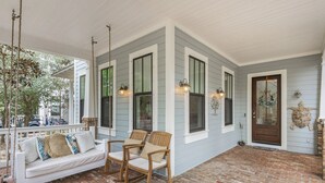 A covered porch with a brick floor, white porch furniture, and a wooden door. - Longleaf Pines by AvantStay | Community Pool (Santa Rosa Beach)