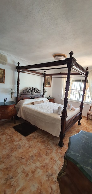 A four-poster bed with a white bedspread, a wooden nightstand with a lamp, and a window with a view of the outside. - House in Fuengirola with private pool, enjoy the relaxation of the... (Fuengirola)