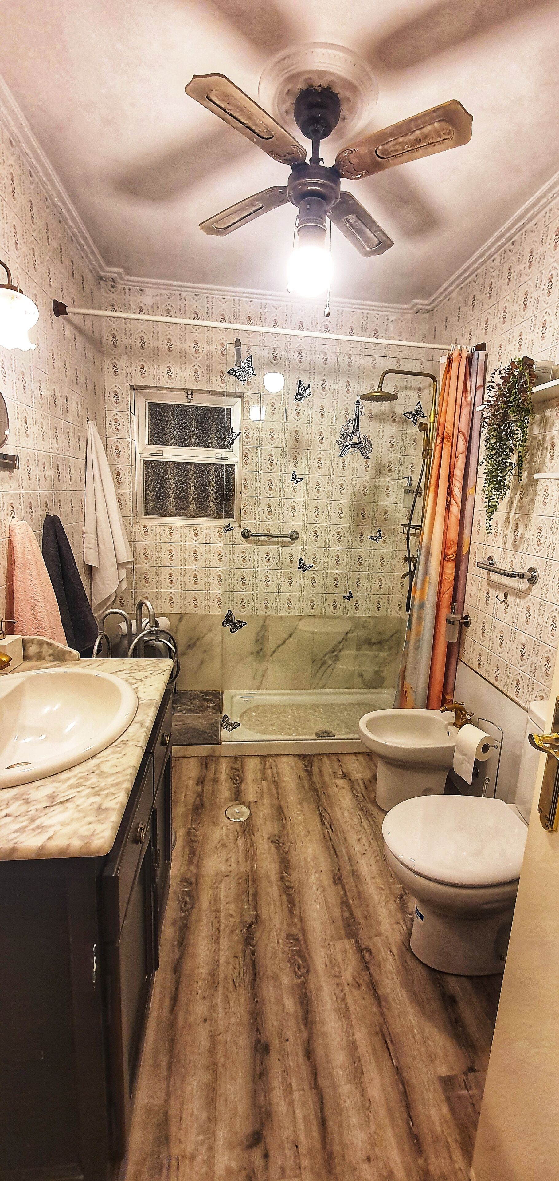 Une salle de bain avec un plancher en bois, un ventilateur de plafond, une douche avec une porte en verre, des toilettes et un lavabo.