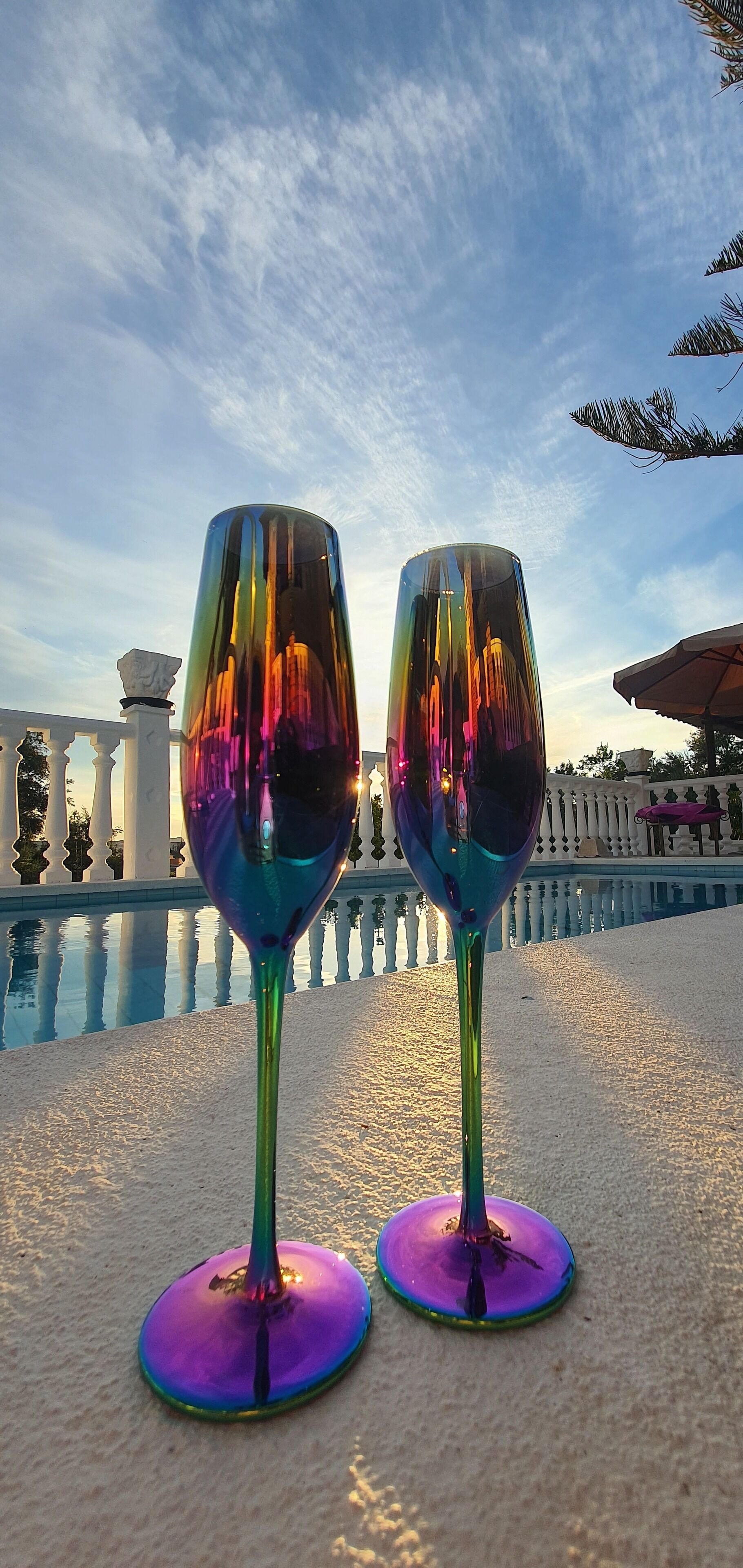 Two wine glasses on a table with a pool and a white fence in the background.