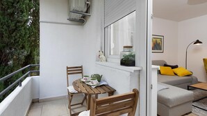 A balcony with a wooden table and chairs, a window with a plant, and a grey sofa with yellow cushions inside a room. - 1 bedroom nice apartment in Pula (Pula)