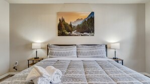 A hotel room with a bed, two bedside lamps, a painting of a mountain landscape, and a folded towel on the bed.