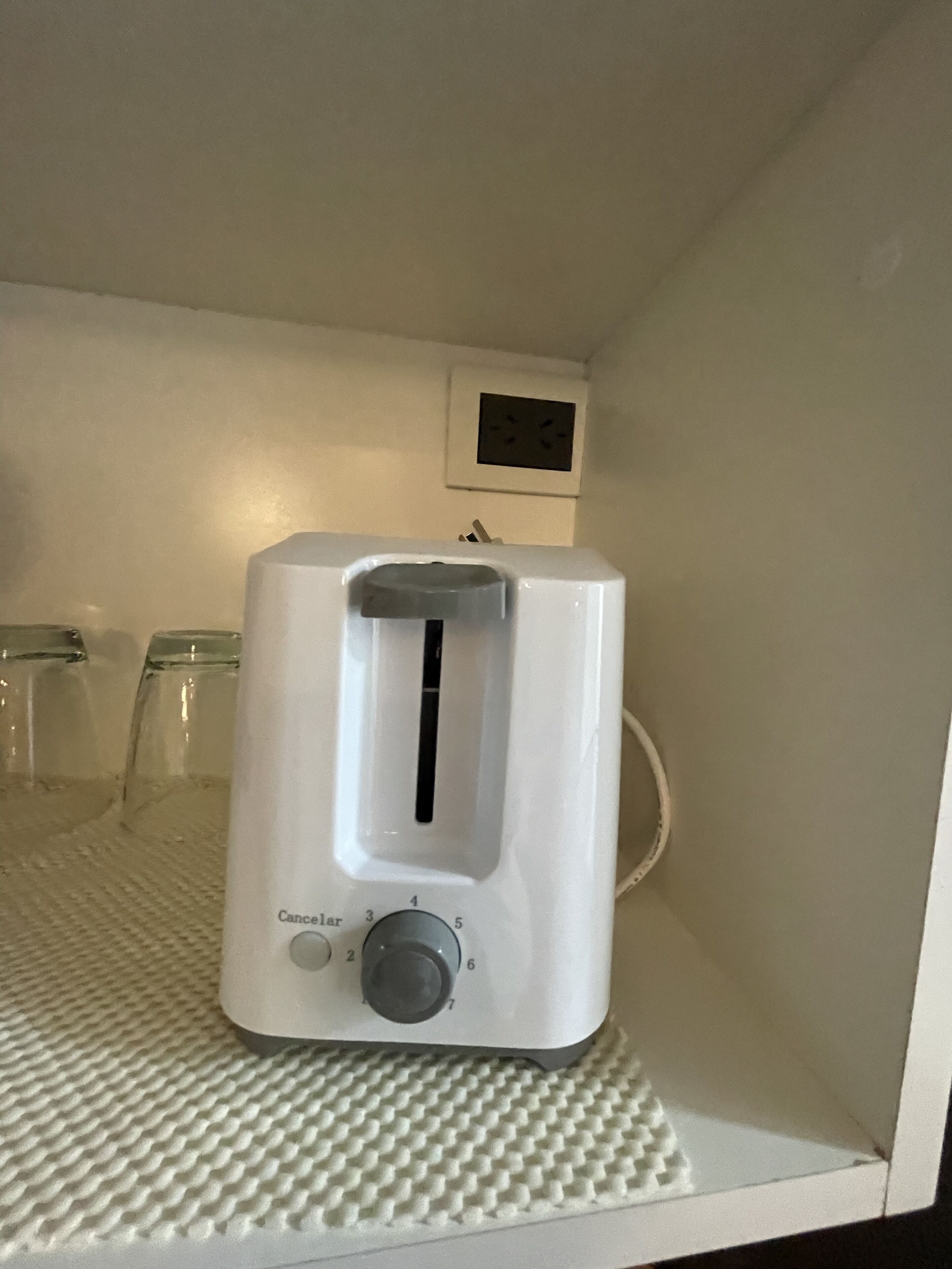 A white toaster on a textured white surface, with a wall-mounted electrical outlet and a small picture frame in the background.