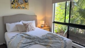 A bedroom with a bed, pillows, a blanket, a nightstand with a lamp, a painting, and a large window with a view of greenery. - Ultimate heaven of comfort and relaxation. Entire house, large private pool.  (Kewarra Beach)