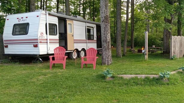 A recreational vehicle (RV) parked in a grassy area with a picnic table and two red chairs nearby. - Russell's Roost Camping , Boating, Fishing ON Martiny Chain of Lakes. (Barryton)