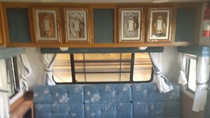 A compact RV interior with a kitchenette, a blue upholstered seating area, and a window with curtains. - Russell's Roost Camping , Boating, Fishing ON Martiny Chain of Lakes. (Barryton)