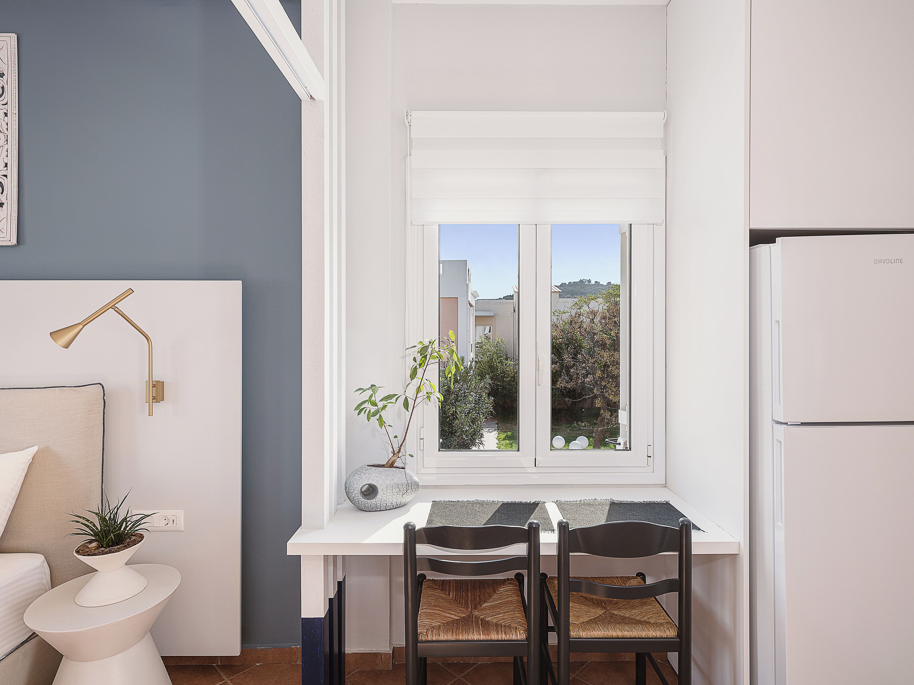 Une cuisine de taille réduite, dotée d’un plan de travail blanc, de deux chaises, d’une plante en pot et d’une fenêtre donnant sur l’extérieur.