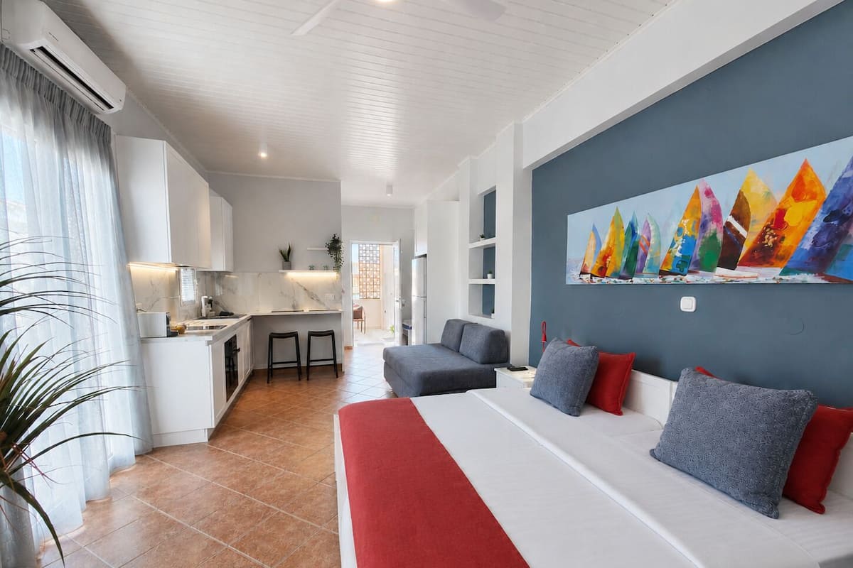 A modern hotel room with a bed, a sofa, a kitchen area, and a dining table.