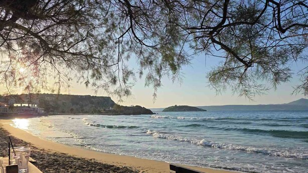 A beach with a wooden bench and a table with glasses and a pitcher, overlooking the sea and a distant island. - Elena SeaFront Luxury Apartments (Apokoronas)