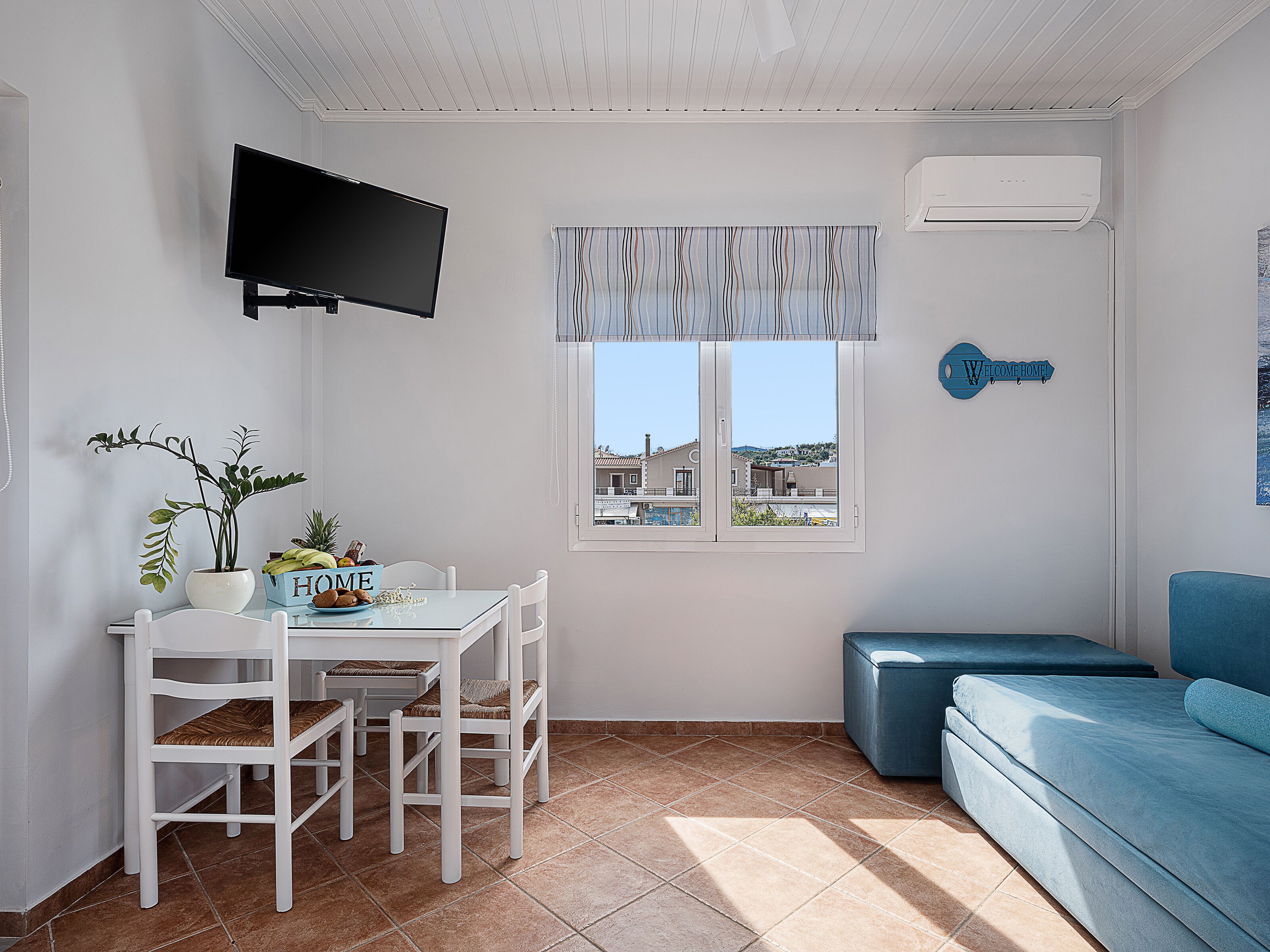 Un espace de vie compact comprenant une table à manger, des chaises, un canapé, un téléviseur fixé au mur, une fenêtre donnant sur des immeubles et un panneau d’accueil.