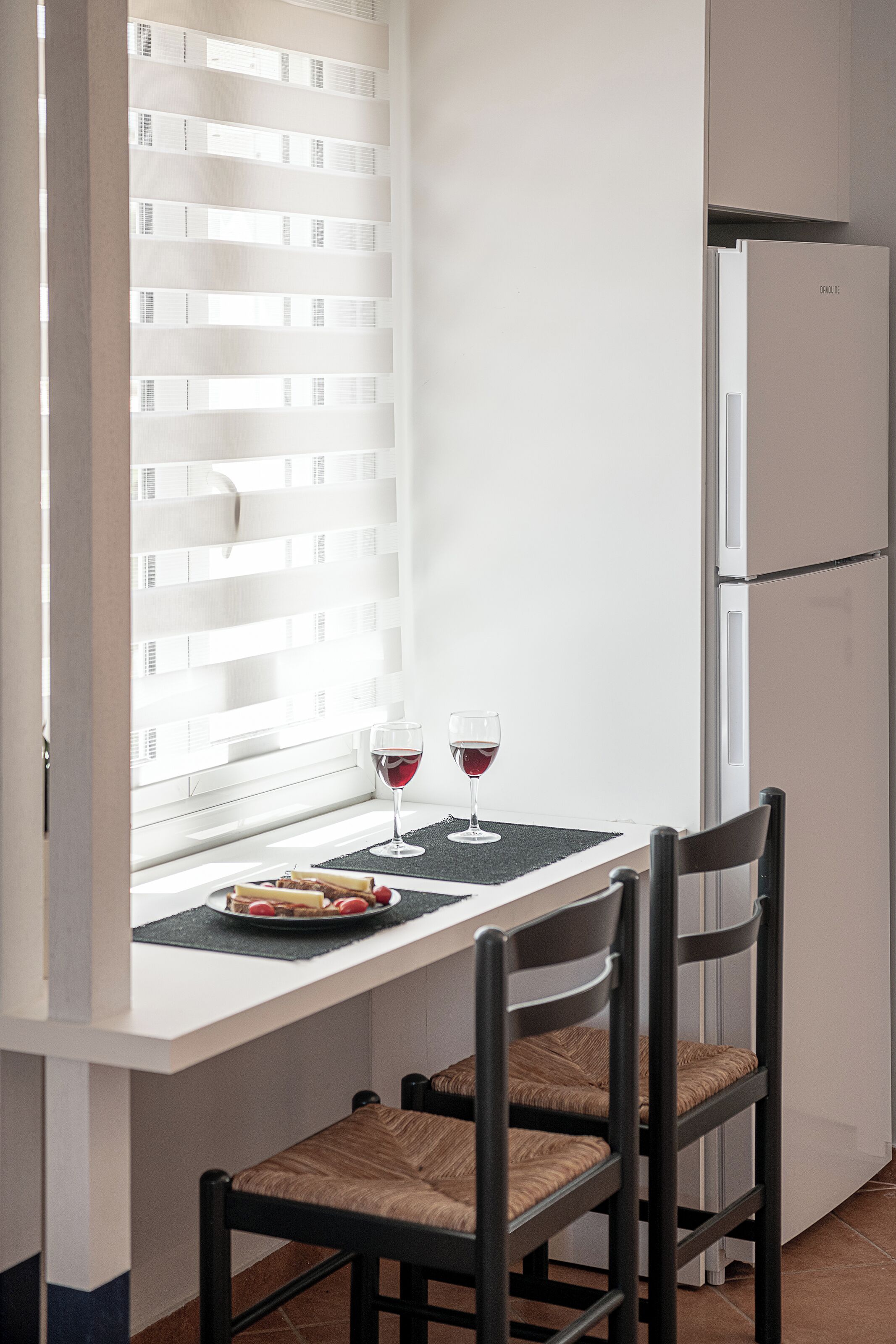 Een kleine keuken met een wit aanrecht, twee stoelen, een koelkast en een raam met jaloezieën.