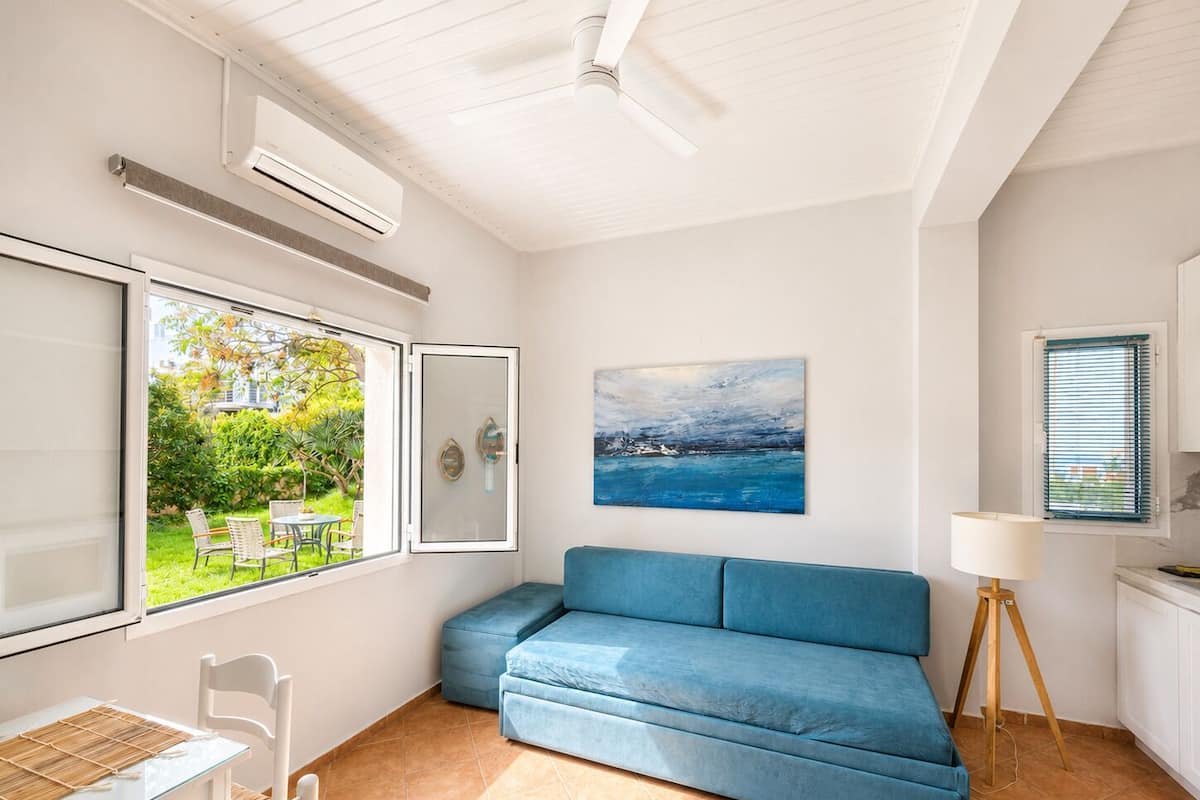 A room with a blue sofa, a painting, a window with blinds, and a ceiling fan.