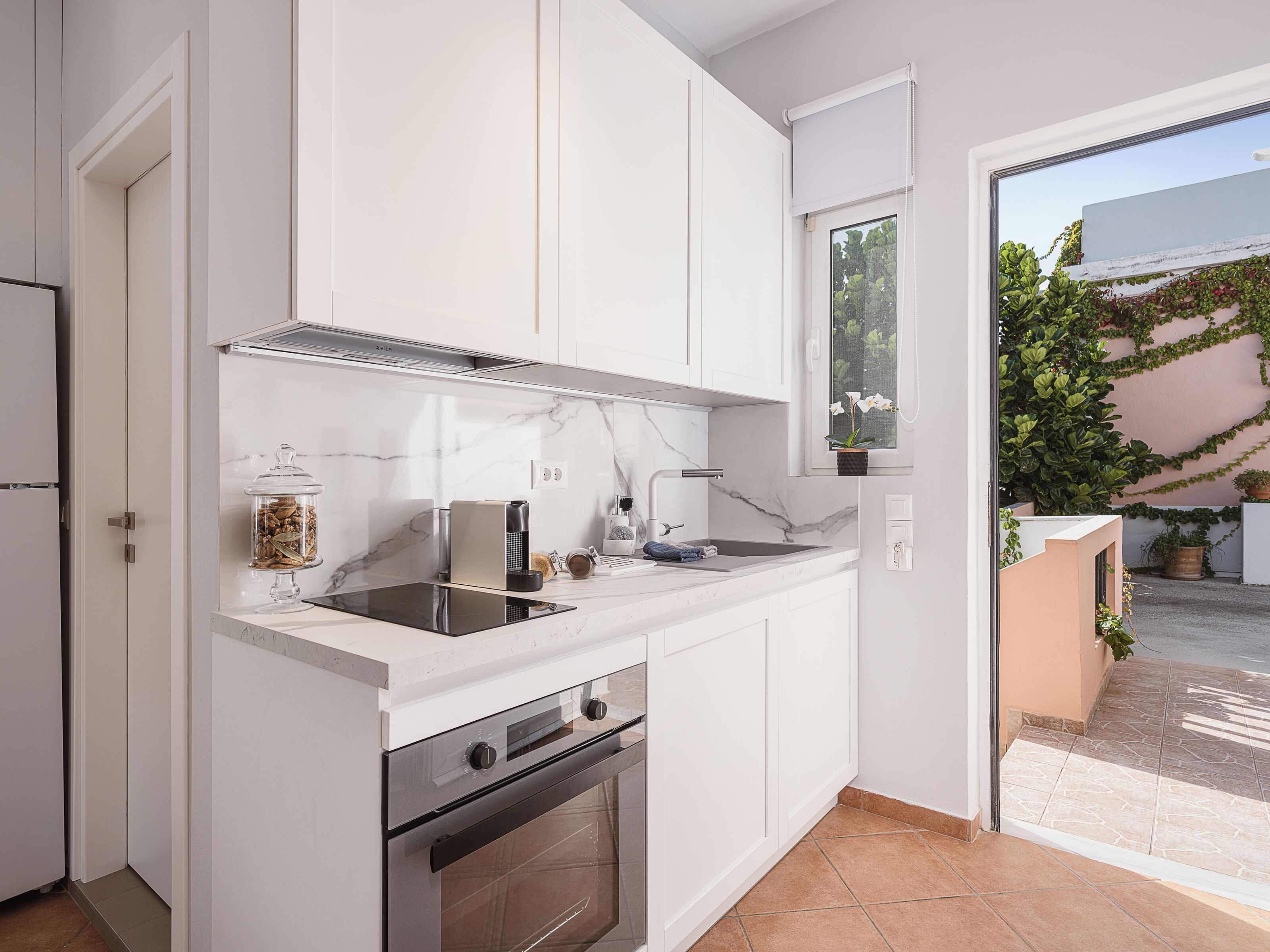 Une cuisine moderne avec des meubles blancs, un four encastré et une fenêtre donnant sur un paysage verdoyant.