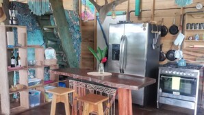 A rustic kitchen with wooden beams, a wooden table with stools, a refrigerator, an oven, and various kitchen utensils hanging on the wall.